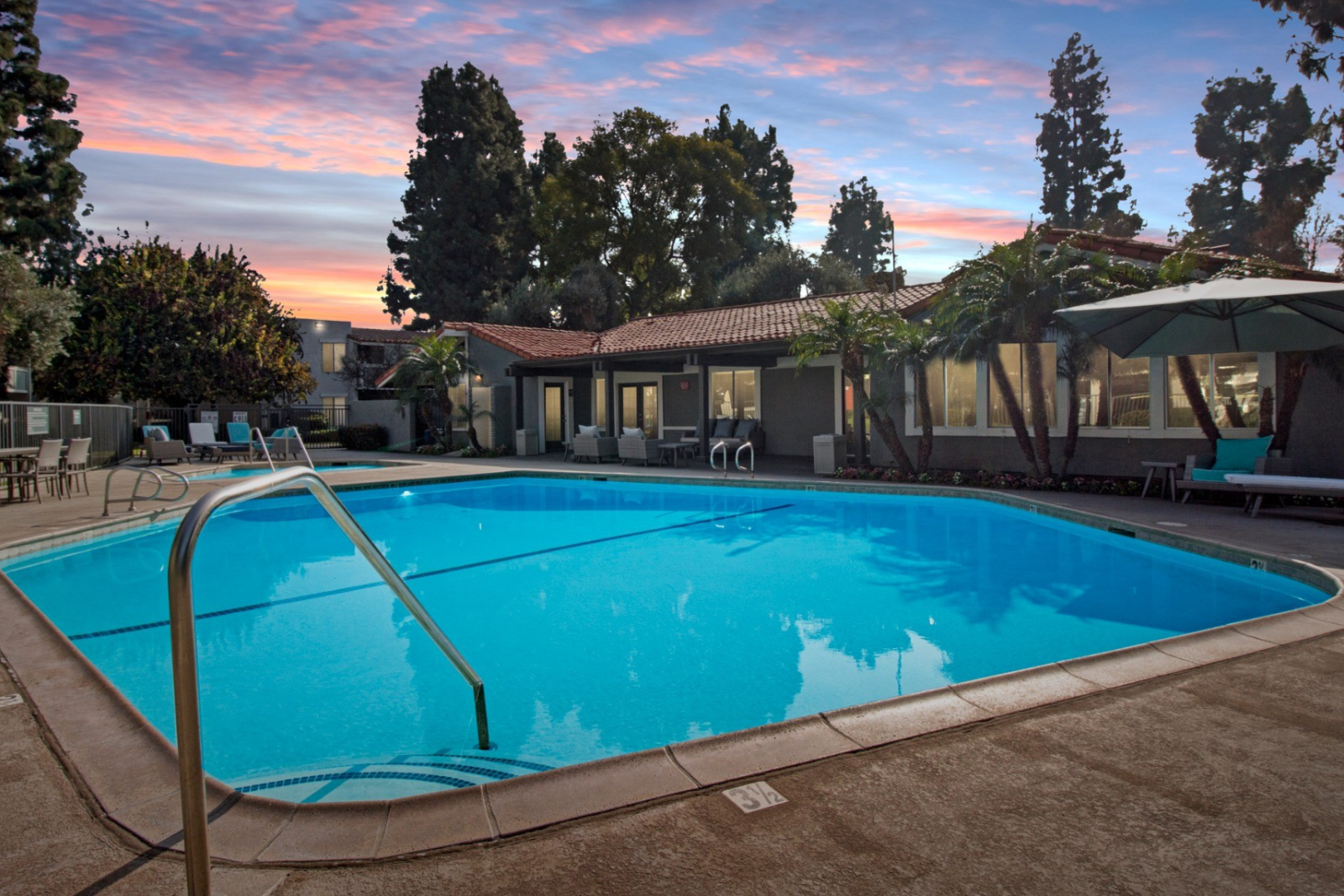 An inviting outdoor pool area at Olive Ridge Apartments, illuminated by the soft glow of the twilight sky. The pool is surrounded by tropical landscaping, including tall palm trees and lush greenery. Lounge chairs and shaded seating areas provide a relaxing environment, with the apartment community's clubhouse in the background.