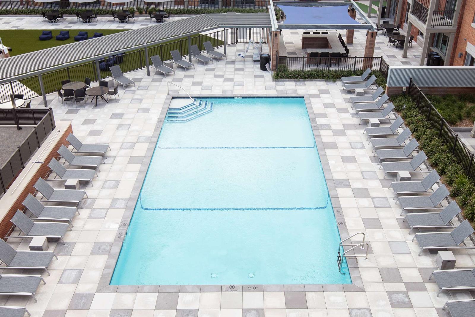 Bright and inviting courtyard pool deck featuring a rectangular swimming pool surrounded by uniform gray tile and sleek lounge chairs neatly arranged on all sides. In the background, a beautifully maintained lawn and shaded cabana area provide extra space for relaxation and social gatherings. The surrounding red-brick apartment buildings with balconies add a warm, urban atmosphere to this serene outdoor amenity space.