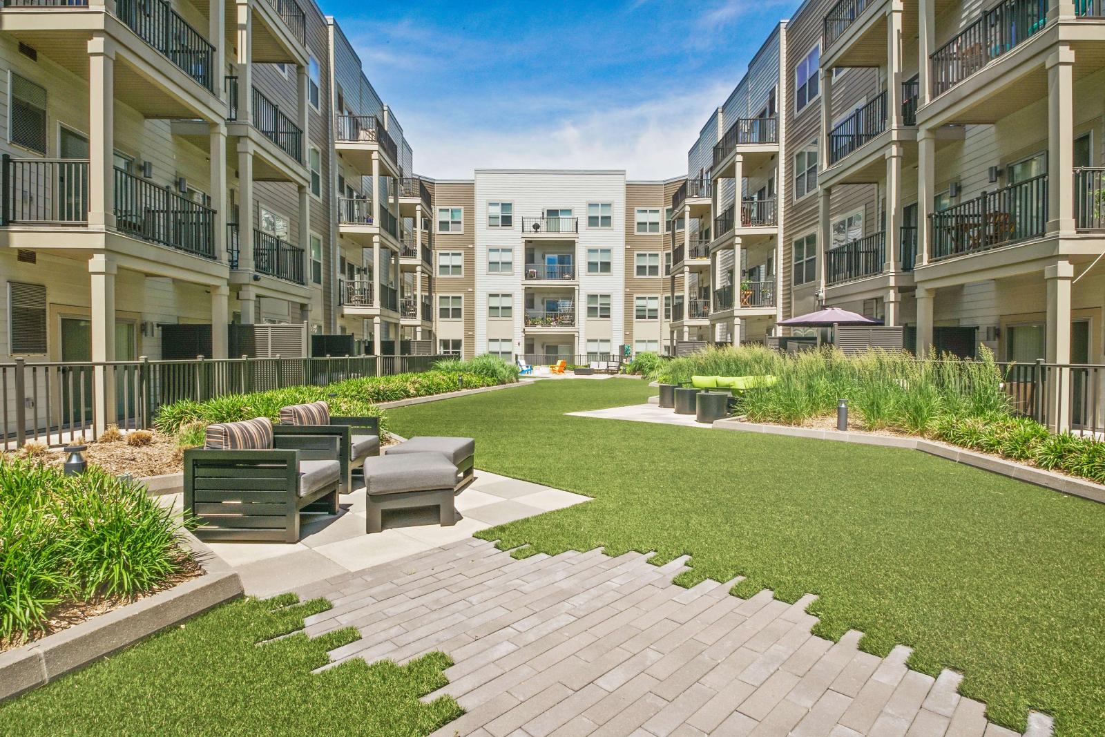 Ground-level view of a vibrant apartment courtyard showcasing artificial turf, modern outdoor seating with cushioned chairs, and well-maintained landscaping. The space is enclosed by four-story residential buildings with balconies, creating a cozy and community-focused atmosphere. The open layout and bright sky provide a welcoming outdoor area perfect for relaxation or social gatherings.