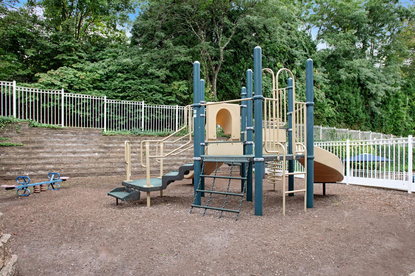 Fenced playground with slides, climbing structures, and mulch ground cover, set against terraced stone steps and tall trees.