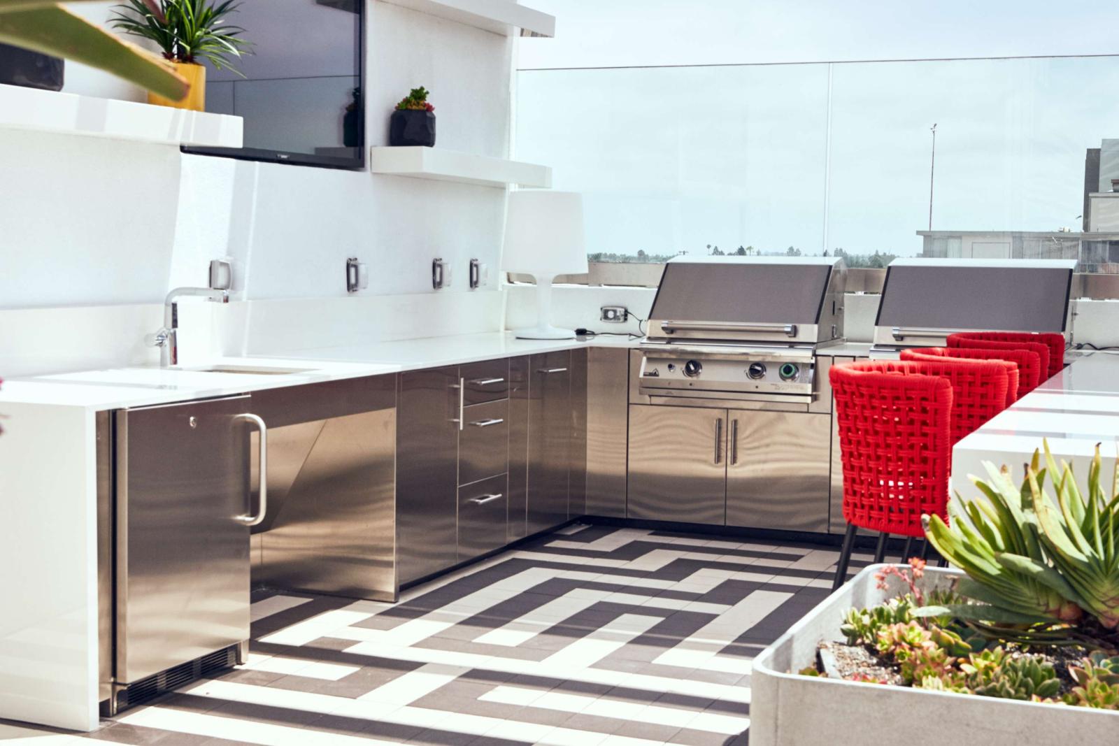 Outdoor rooftop kitchen area with stainless steel appliances, red barstools, and modern tile flooring.