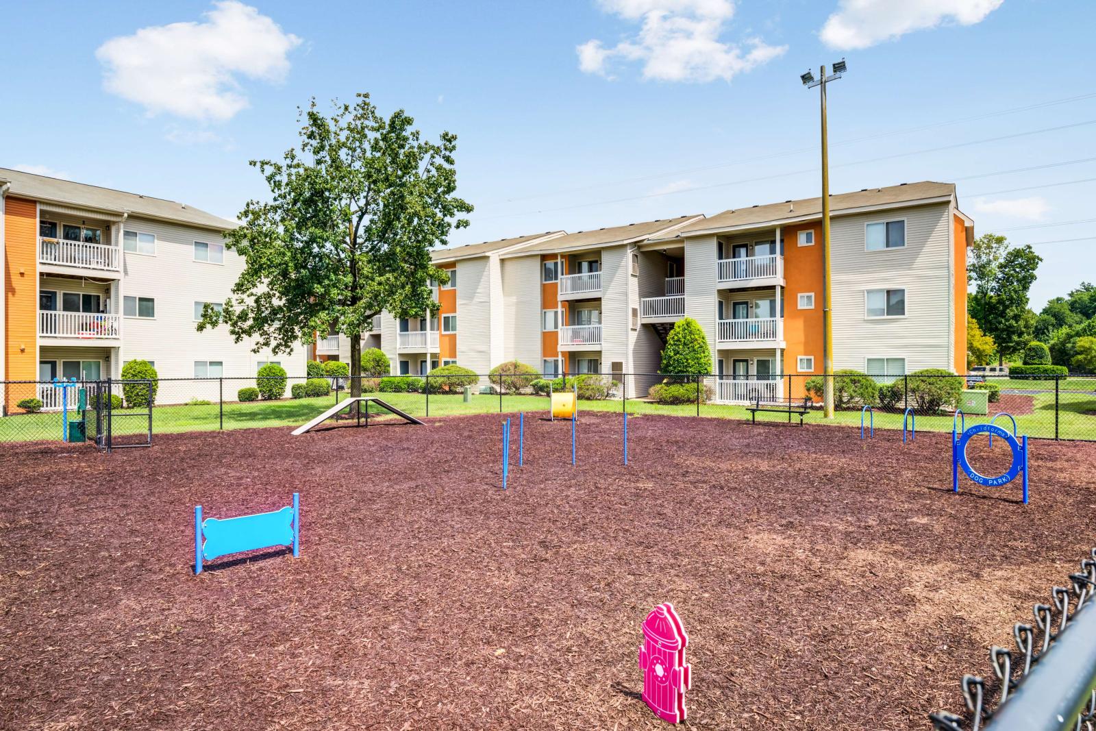 Fenced-in dog park at The Samuel with agility equipment and nearby apartment buildings in the background