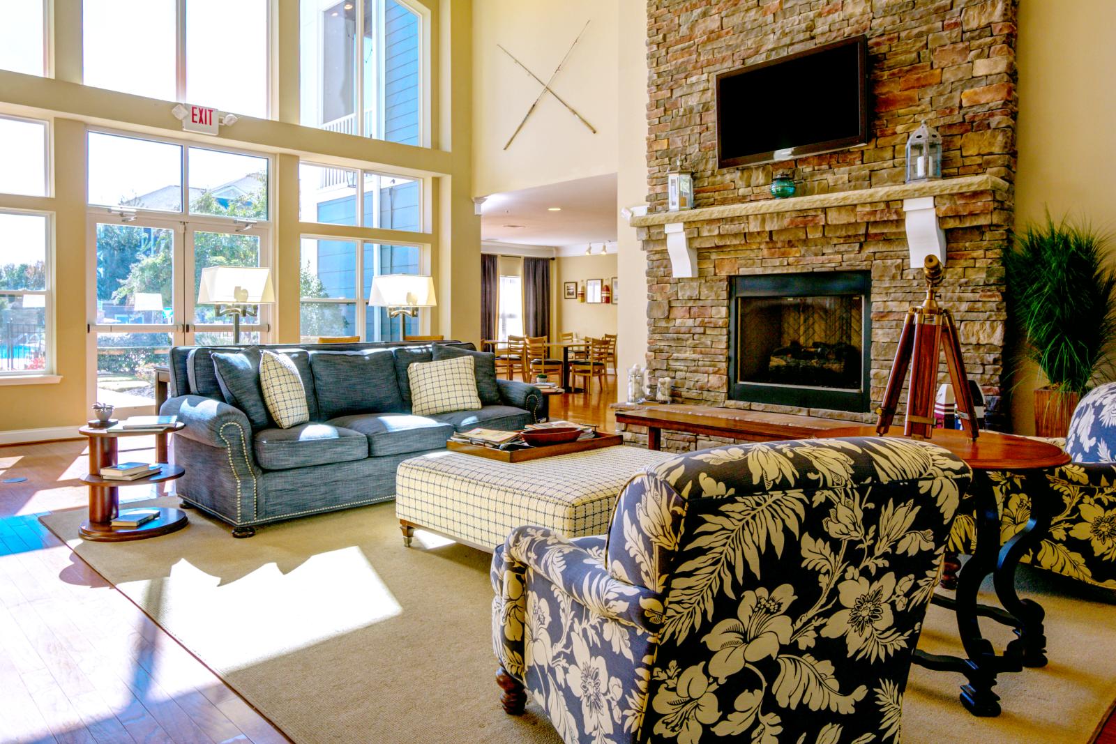 Bright community lounge with cozy furniture, a stone fireplace, and natural light from floor-to-ceiling windows