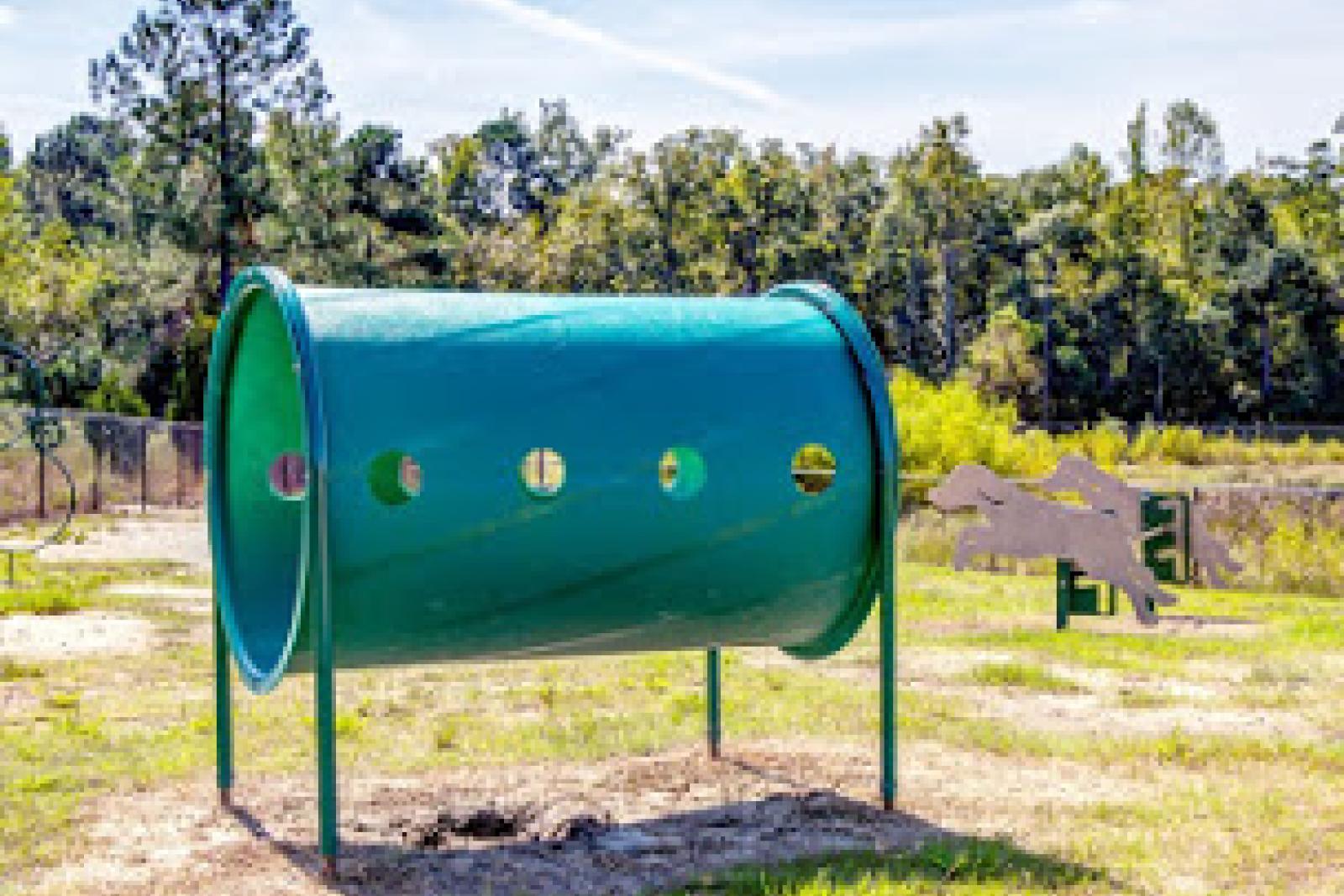 Green agility tunnel in fenced dog park area, surrounded by grass and mature trees.