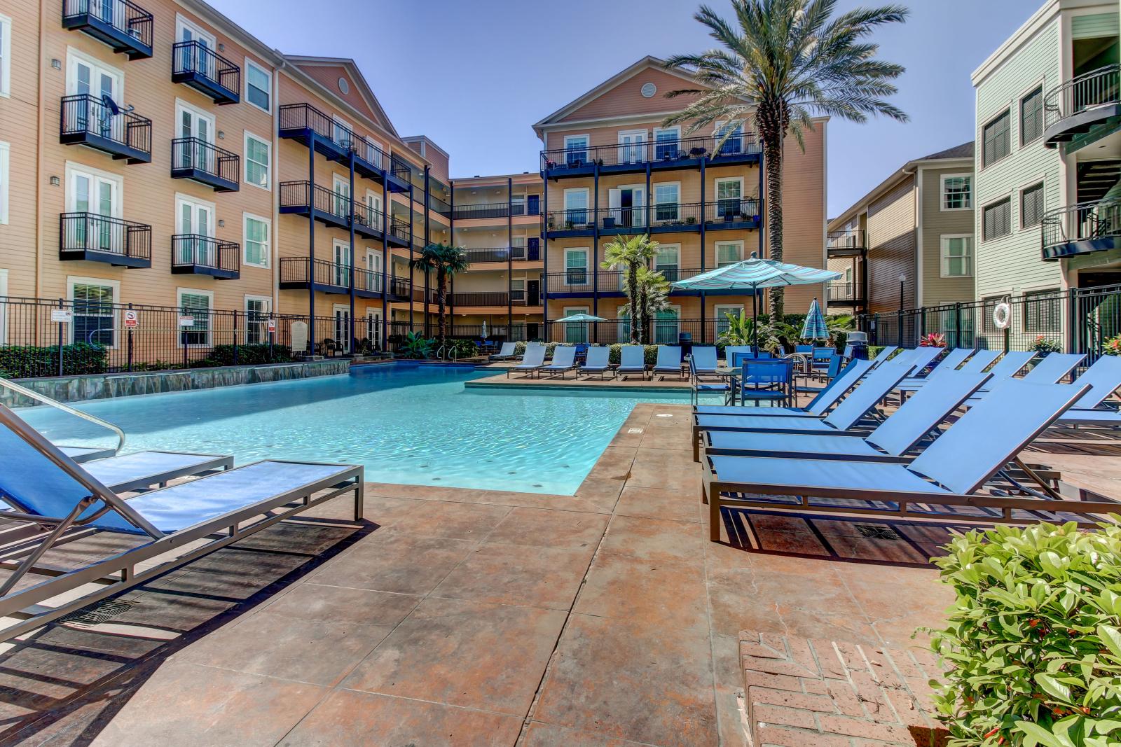 Outdoor pool area with plenty of seating and palm trees, surrounded by colorful apartment buildings at The Saulet Apartments.