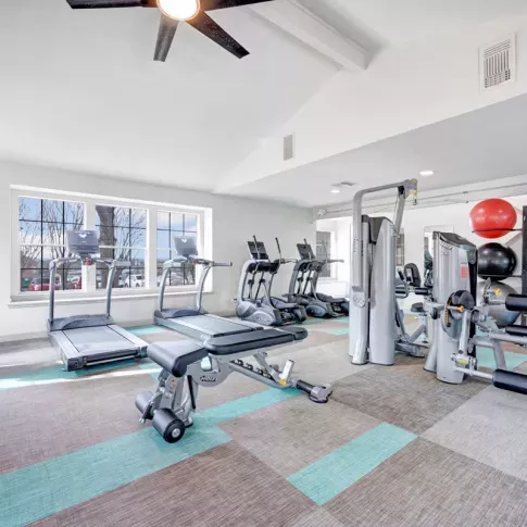 Bright and spacious fitness center equipped with modern exercise machines, including treadmills, ellipticals, and strength-training equipment. The space features large windows, colorful flooring, and ceiling fans, creating an inviting and energizing workout environment.