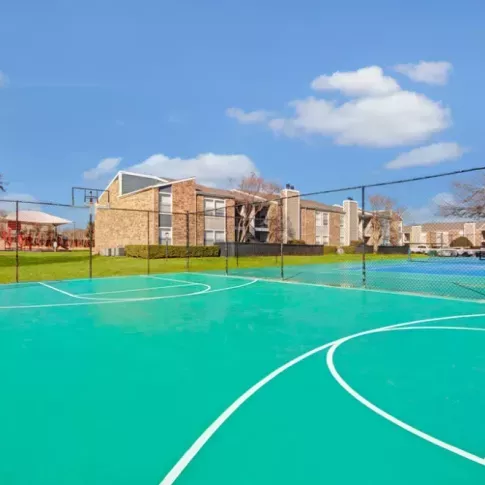 Outdoor basketball court with a vibrant green surface, surrounded by apartment buildings and green lawns.