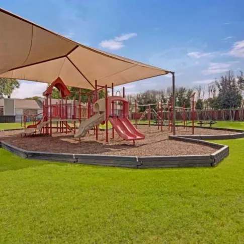 A vibrant playground with red slides and climbing equipment shaded by a large canopy in a green park setting.
