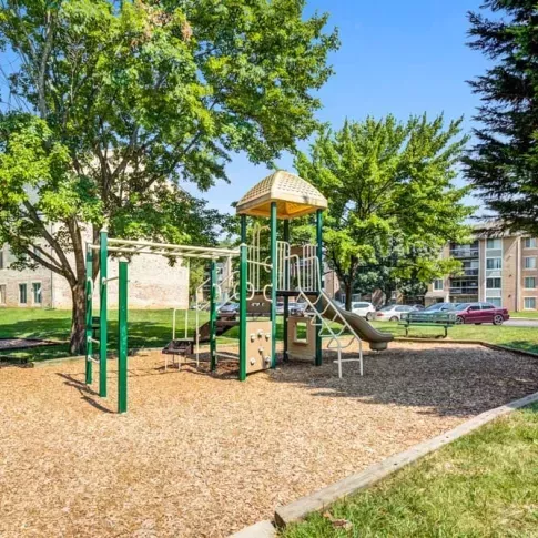 A vibrant playground with slides and climbing structures surrounded by mature trees and apartment buildings in the background.