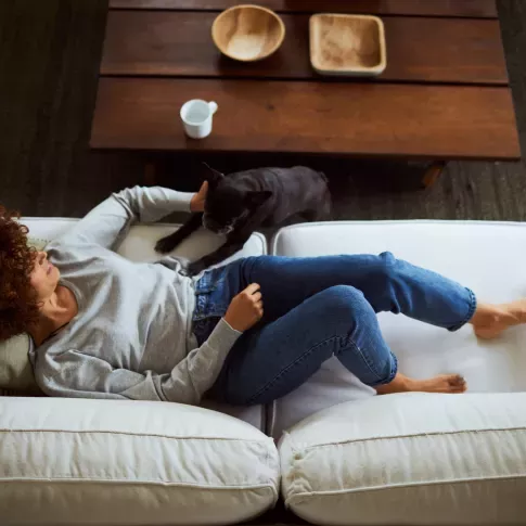A woman lounging on a white couch is petting her black dog, with a wooden coffee table and cozy decor in the background.