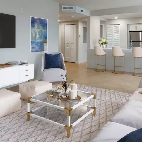 A bright and modern living room with white and gold decor, a mounted TV, and a view into an open-concept kitchen with a bar area.