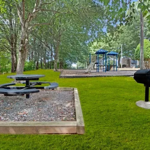A picnic area with a circular table and a grill adjacent to a playground in a wooded setting.