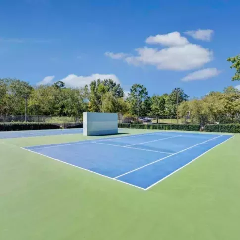 A vibrant blue tennis court sits under a clear blue sky, surrounded by lush greenery.
