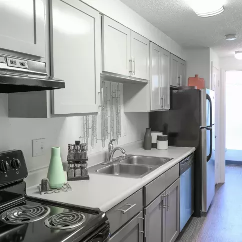 A modern galley kitchen with white cabinets, black appliances, a double sink, and a sleek countertop, leading to a well-lit hallway.