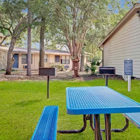 A vibrant outdoor picnic area featuring a blue metal table and a grill, surrounded by lush green grass and mature trees. This image showcases the community amenities and inviting outdoor spaces at Broadview Oaks Apartments.