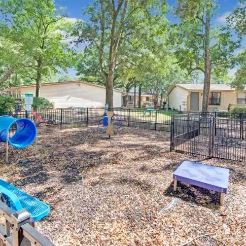 A fenced dog park featuring a blue tunnel slide, climbing equipment, and shaded areas, surrounded by trees and nearby apartment buildings. This image highlights the family-friendly amenities available at Broadview Oaks Apartments.