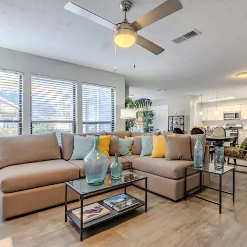 A spacious living room featuring a large sectional sofa with colorful throw pillows, a ceiling fan, and large windows letting in ample natural light.