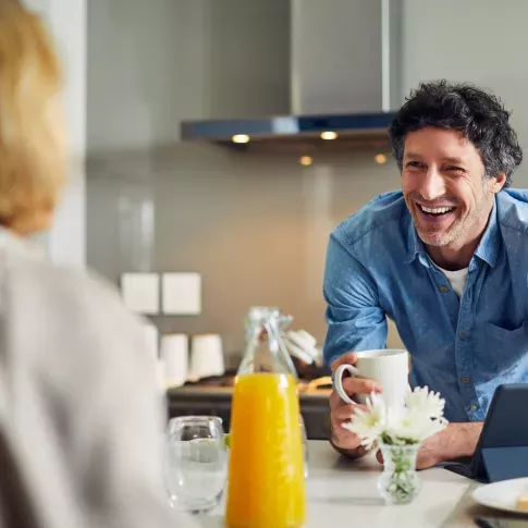 A man in a blue shirt smiles warmly while holding a coffee mug, with a breakfast setup of orange juice, flowers, and a tablet on the table.