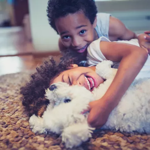 Two children happily cuddle and play with a fluffy white dog on a cozy rug in a brightly lit home.