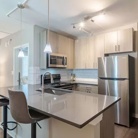 A modern kitchen with sleek stainless steel appliances, including a refrigerator and microwave, set against a light-colored cabinet backdrop. The island features bar stools, and the space is illuminated with stylish pendant lighting