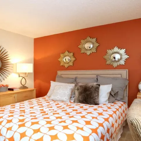  A stylish bedroom at Norcross Crossings Apartments features a bold orange accent wall, elegant decor with sunburst mirrors, and cozy furnishings for a warm and inviting atmosphere.
