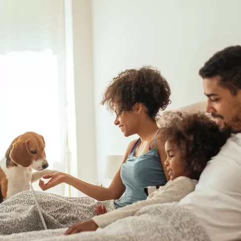 A family of three, including a young child, enjoys a relaxing moment in bed with their pet dog, smiling and engaging with each other in a cozy setting.