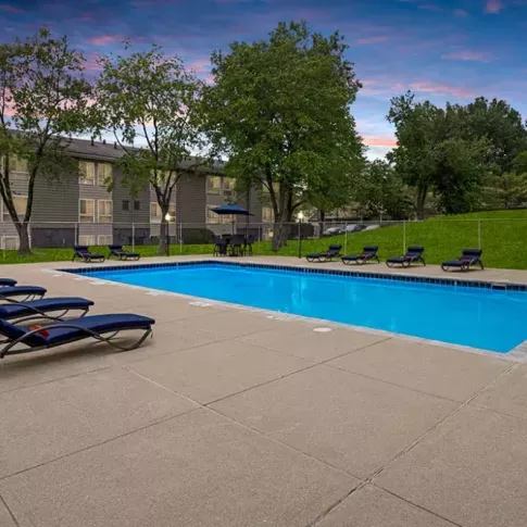 Outdoor swimming pool surrounded by lounge chairs, with a sunset sky and a grassy area in the background.