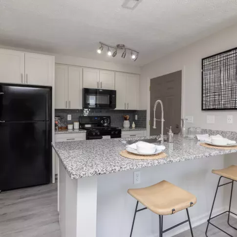 Modern kitchen with sleek granite countertops, bar seating, stainless steel sink, black appliances, and contemporary artwork, offering a stylish and functional living space in the apartment community.