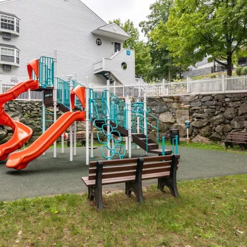 A colorful playground featuring orange slides and turquoise climbing structures, surrounded by benches and set against a backdrop of stone walls and residential buildings.