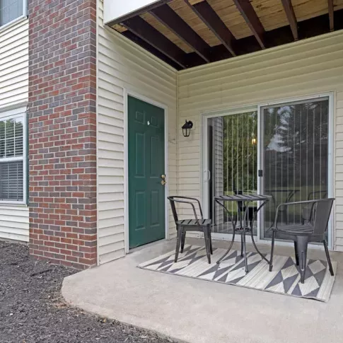 A cozy outdoor patio space with a small table, two chairs, and a patterned rug, overlooking a green yard.