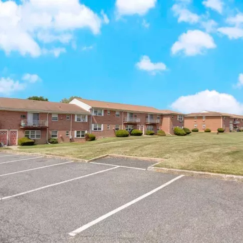 Wide parking lot and brick apartment buildings with balconies at Washington Gardens Apartments in Washington, NJ, under a bright blue sky.