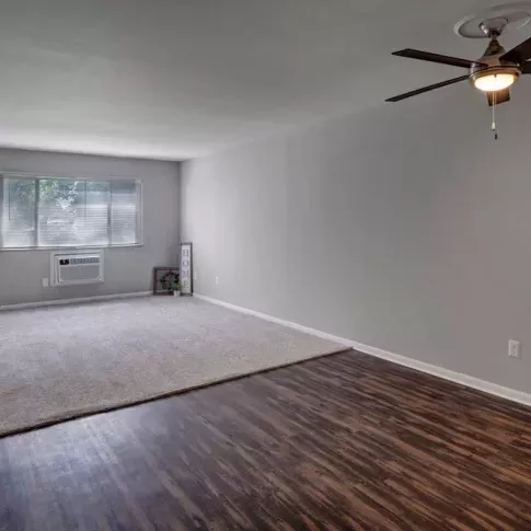 A spacious living room with wood-style flooring, a ceiling fan, and natural light from large windows at Yearling Plaza Apartments in Columbus, OH.