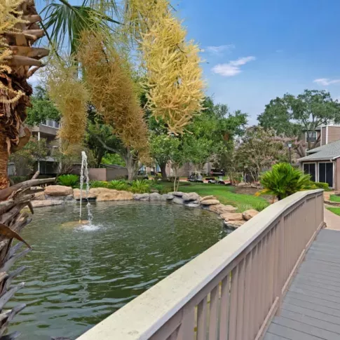A picturesque outdoor scene with a charming wooden bridge over a tranquil pond, surrounded by lush greenery, blooming flowers, and a decorative fountain.