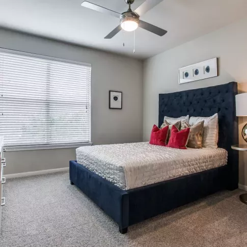 A cozy bedroom with a tufted navy headboard, red accent pillows, and a ceiling fan, creating a relaxing space.