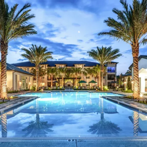 A beautifully illuminated pool area at dusk, reflecting palm trees and the surrounding modern apartments.