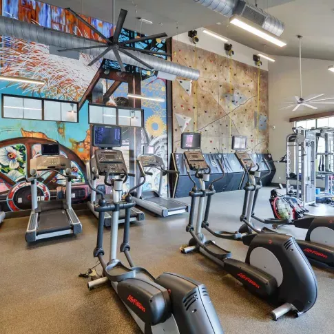 "Modern fitness center at Riverworks Luxury Apartments with treadmills, ellipticals, and vibrant wall art alongside a rock climbing wall."
