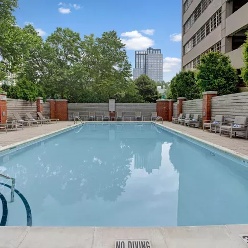 A spacious outdoor pool area with modern seating, trees, and the apartment buildings in the background.
