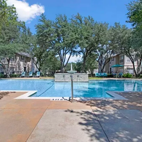 Sparkling pool with a stone water fountain, surrounded by mature trees and sun loungers.
