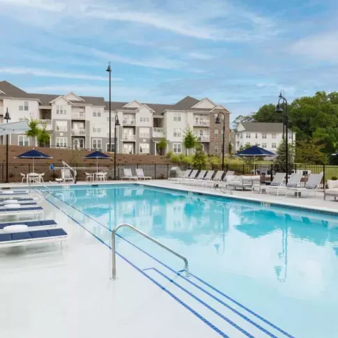 A sparkling outdoor pool surrounded by elegant lounge seating and shaded cabanas, set against a backdrop of luxury apartment buildings and green space.