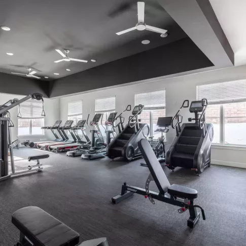 Spacious gym area featuring treadmills, stair climbers, and cardio equipment with large windows offering natural light.
