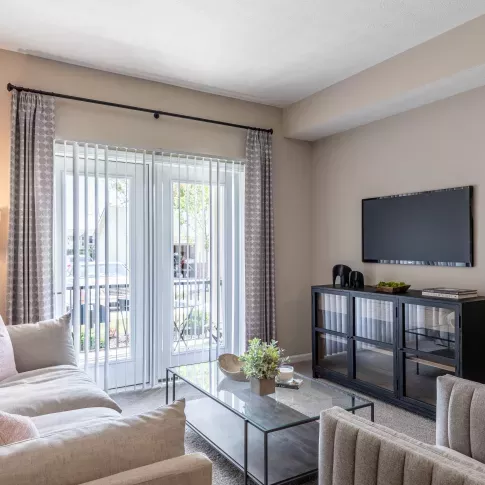A cozy living room at Victoria Manor Luxury Apartments with soft beige furnishings, large windows, and a wall-mounted TV.