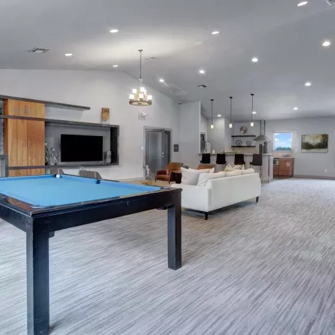 Modern clubhouse interior featuring a blue felt pool table, cozy seating area with a white couch, bar stools at a kitchen counter, and large windows allowing natural light.