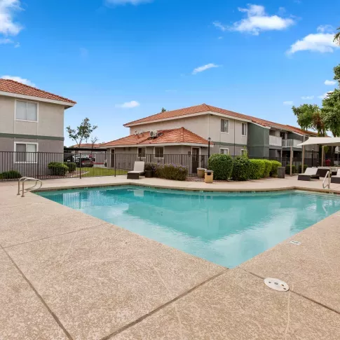 Outdoor swimming pool surrounded by a spacious sundeck with modern lounge chairs, set against a backdrop of two-story apartment buildings and palm trees.
