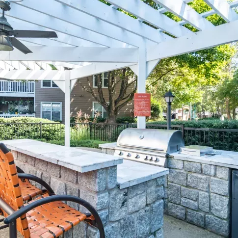 Outdoor grilling station under a white pergola with bar seating and ceiling fans, surrounded by lush greenery.