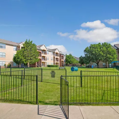 Gated entrance to a spacious dog park with agility tunnel and hurdles, surrounded by grassy lawns and apartment buildings.