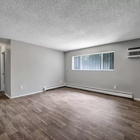 Spacious living room with wood-style flooring, neutral walls, and a large horizontal window with vertical blinds and an in-wall AC unit.