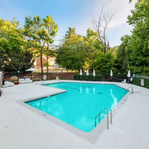 Resort-style outdoor swimming pool surrounded by white lounge chairs, umbrellas, and lush greenery, offering a peaceful and inviting setting for relaxation.