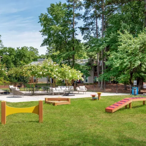 Scenic outdoor pet park and seating area with agility equipment, shaded lounge furniture, and mature trees providing a peaceful, community-friendly space.
