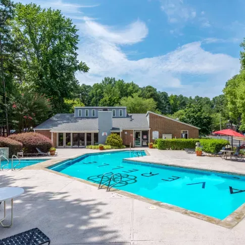Expansive outdoor pool with crystal-clear water, surrounded by greenery and a brick clubhouse, with plenty of lounge seating and red umbrellas for shaded relaxation.