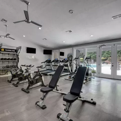 Well-equipped fitness center with treadmills, ellipticals, spin bikes, free weights, and functional training equipment, featuring modern flooring and large windows overlooking the pool area.